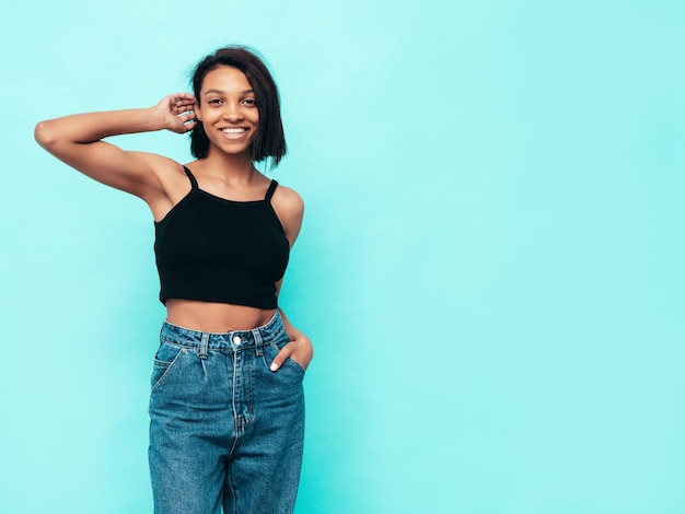 Portrait de jeune belle femme noire Modèle souriant vêtu d'un jean d'été et de hauts vêtements noirs Sexy femme insouciante posant près du mur bleu en studio Bronzé et joyeux