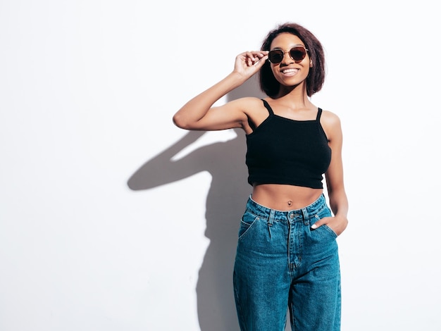 Portrait de jeune belle femme noire Modèle souriant vêtu d'un jean d'été et de hauts vêtements noirs Sexy femme insouciante posant près d'un mur blanc en studio Bronzé et joyeux