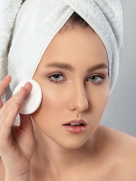 Portrait de jeune belle femme avec une serviette sur les cheveux nettoyant son visage avec un tampon de coton cosmétique Nettoyage de routine du matin visage peau parfaite SPA thérapie soins de la peau concept de cosmétologie et de maquillage