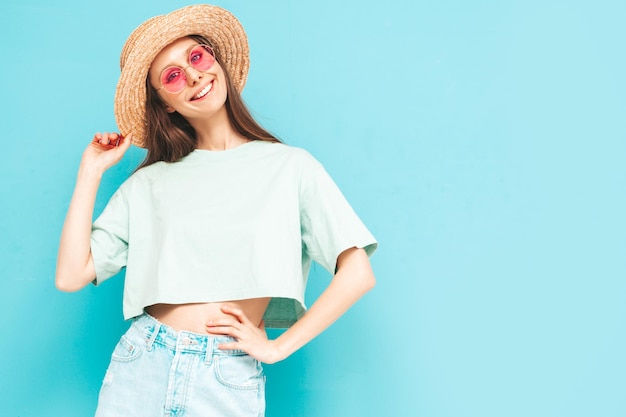 Portrait de jeune belle femme souriante en jeans d'été à la mode jupe femme insouciante posant près du mur bleu en studio Modèle positif s'amuser à l'intérieur en lunettes de soleil Enthousiaste et heureux Au chapeau