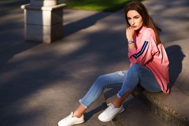 Portrait de jeune et belle femme en vêtements décontractés dans la rue. vêtu d'une chemise rose et d'un jean. concept printemps / été. temps de repos. fille aux yeux bleus. Profitez du coucher de soleil dans la rue