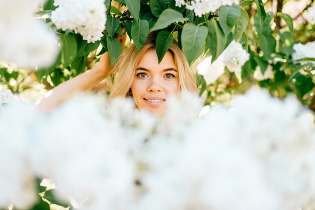 Portrait de jeune belle heureuse joyeuse souriante fille blonde positive regardant à travers les branches avec des fleurs blanches dans le parc fleuri d'été.