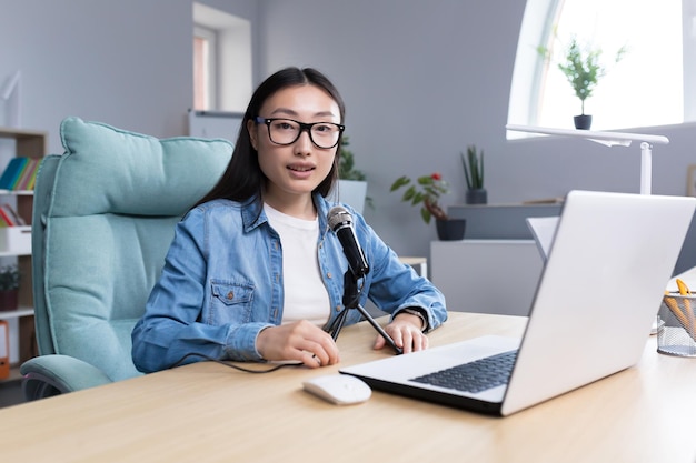 Portrait d'une jeune blogueuse asiatique belle et réussie souriante et regardant la caméra travaillant dans un bureau de studio enregistrant un podcast audio à l'aide d'un microphone professionnel