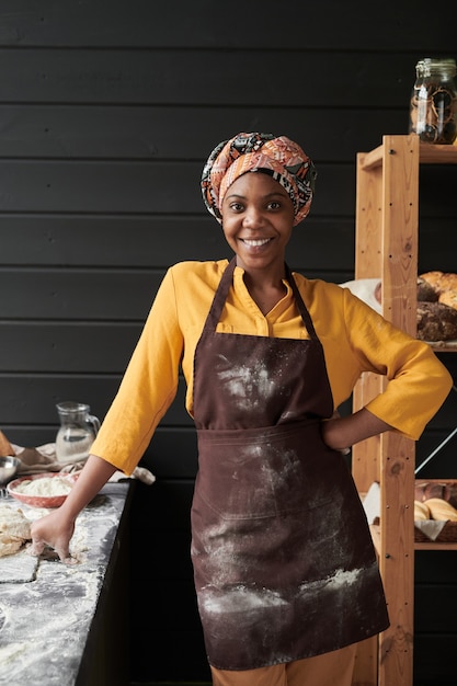 Portrait de jeune boulanger africain en tablier souriant à la caméra en se tenant debout dans la cuisine