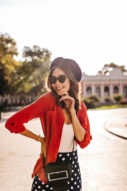 Portrait de jeune brune joyeuse en lunettes de soleil béret français haut blanc chemise rouge et jupe à pois souriant et posant à l'extérieur en regardant ailleurs Ville chaude et ensoleillée