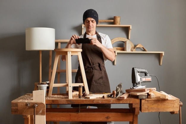 Portrait d'un jeune charpentier portant un chapeau noir et un tablier marron posant dans son atelier de menuiserie prenant une photo au téléphone de son produit fabriquant des chaises en bois