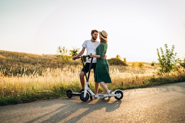 Portrait de jeune couple au coucher du soleil avec un beau paysage naturel. Marcher sur des scooters électriques à la campagne