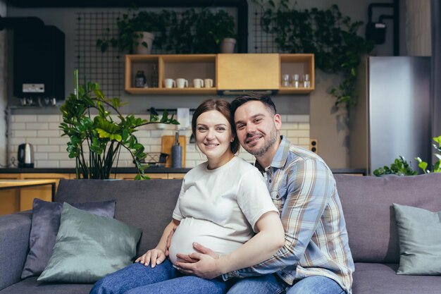 Portrait de jeune couple de famille aimante homme étreignant une femme enceinte souriant et regardant la caméra à la maison assis sur un canapé