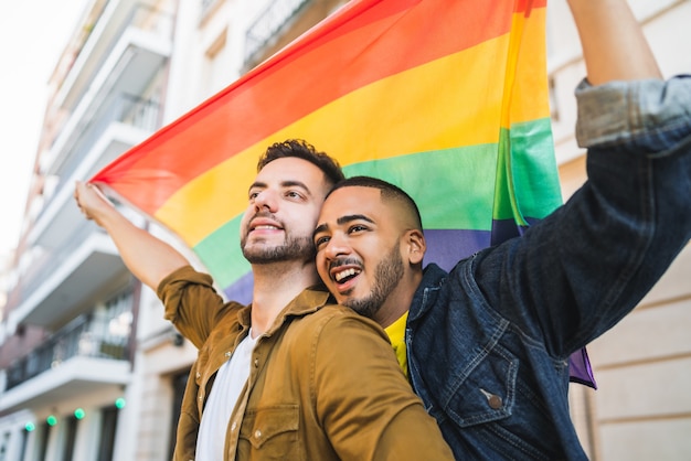Portrait de jeune couple gay embrassant et montrant leur amour avec le drapeau arc-en-ciel dans la rue