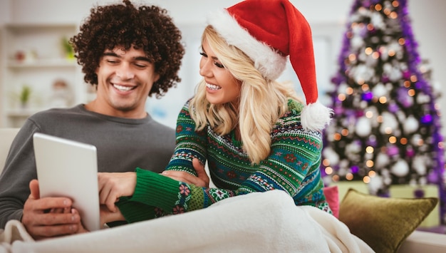 Portrait d'un jeune couple heureux célébrant Noël à la maison, se relaxant et regardant quelque chose d'amusant sur une tablette.