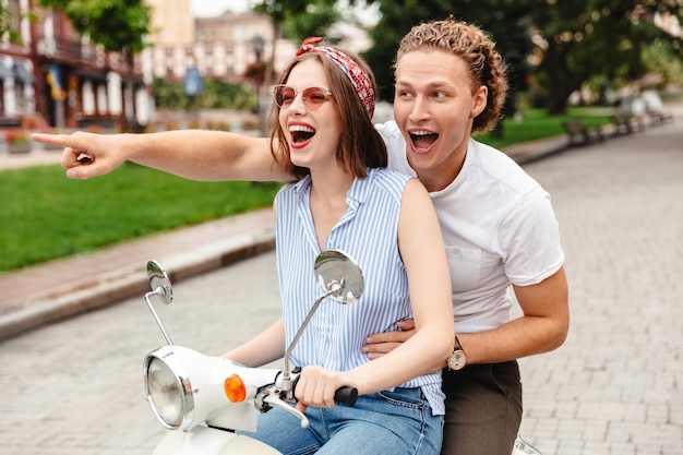 Portrait d'un jeune couple joyeux à cheval sur une moto ensemble à la rue de la ville, doigt pointé