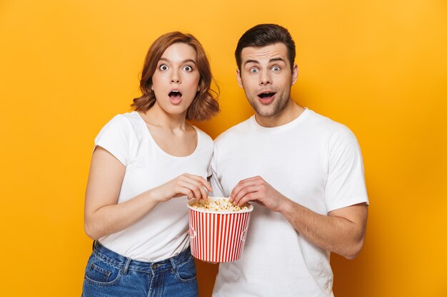 Portrait d'un jeune couple joyeux isolé sur un mur jaune, mangeant du pop-corn