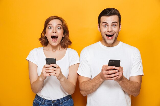 Portrait d'un jeune couple joyeux isolé sur un mur jaune, utilisant des téléphones portables