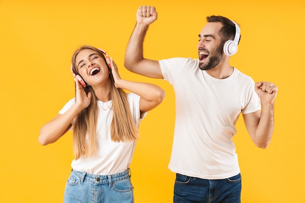 Portrait d'un jeune couple joyeux et séduisant portant des vêtements décontractés, isolé sur un mur jaune, écoutant de la musique avec des écouteurs, dansant