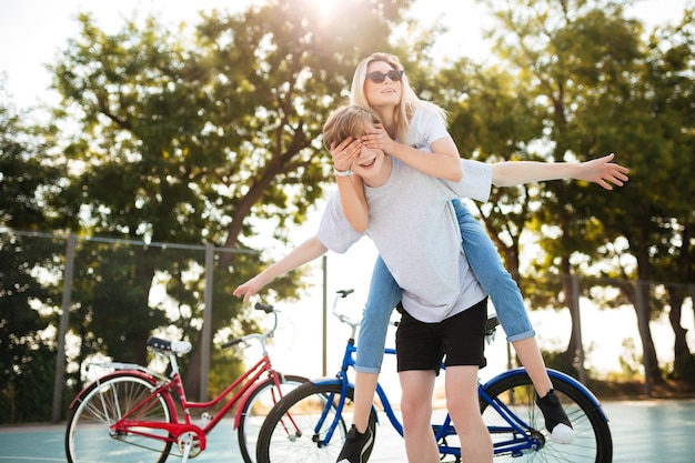 Portrait de jeune couple s'amusant ensemble dans le parc avec des vélos en arrière-plan Joyeux garçon jouant avec une belle fille à lunettes de soleil et montrant le geste des ailes d'avion pendant qu'elle lui fermait les yeux