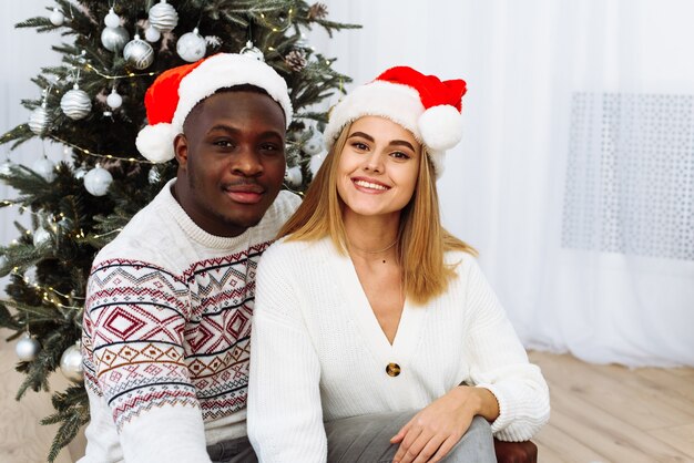 Portrait de jeune couple souriant s'asseoir près de l'arbre de Noël pour célébrer le Nouvel An ensemble. Un homme et une femme heureux profitent de la célébration des vacances d'hiver près d'un sapin décoré.