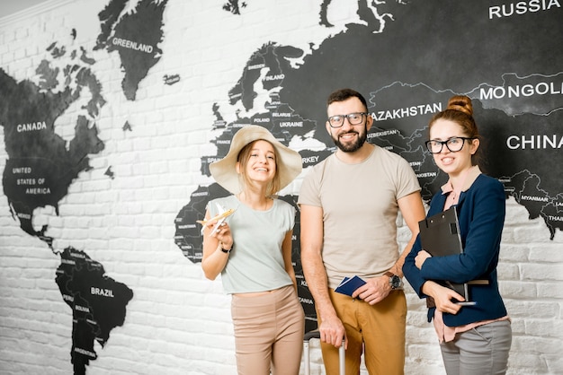 Portrait d'un jeune couple de voyageurs choisissant une bande d'été debout avec un agent de voyages sur fond de carte du monde