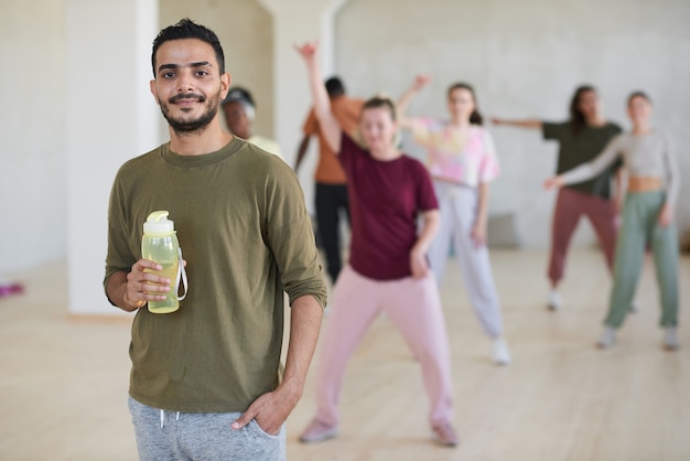 Portrait de jeune danseur avec une bouteille d'eau souriant à la caméra en se tenant debout dans le studio de danse et la formation