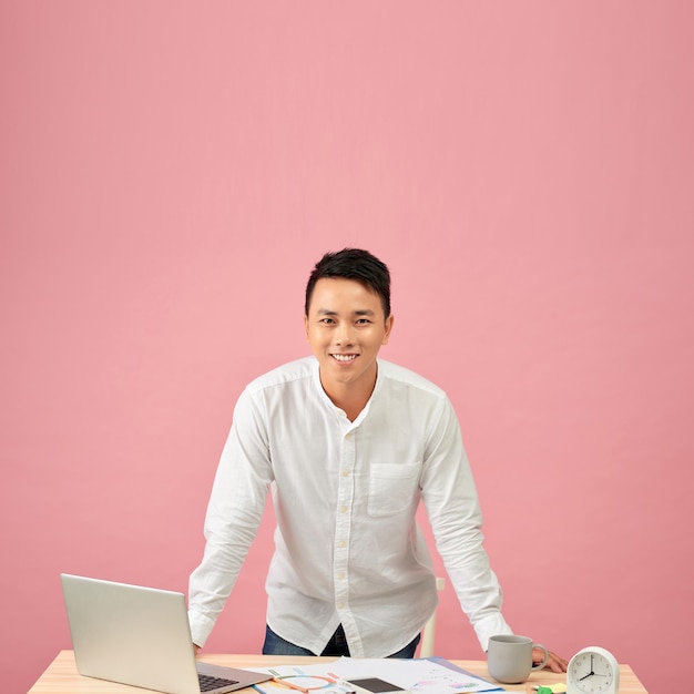 Portrait d'un jeune designer asiatique souriant travaillant sur un ordinateur portable tout en s'appuyant sur son bureau