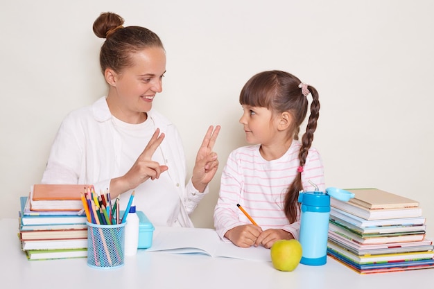 Portrait d'un jeune enseignant tuteur aide un petit élève avec des mathématiques expliquant la tâche via des doigts souriant posant isolé sur fond blanc assis entouré de livres