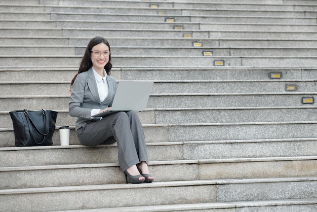 Portrait d'une jeune entrepreneure positive assise sur des marches à côté de son sac et d'une tasse de café et travaillant sur un ordinateur portable