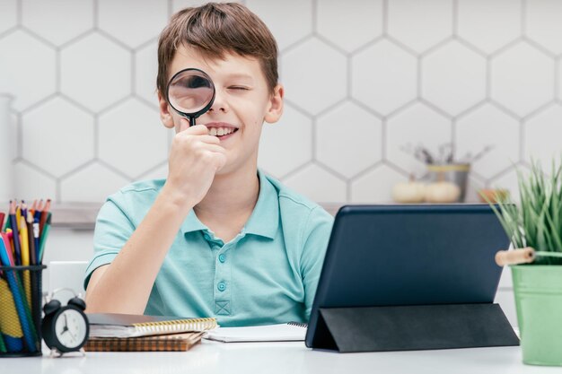 Portrait d'un jeune étudiant souriant assis au bureau avec une tablette numérique de crayons regardant à travers une loupe