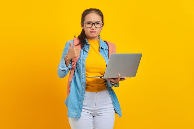 Portrait d'une jeune étudiante asiatique agacée dans des vêtements décontractés avec sac à dos à l'aide d'un ordinateur portable et montrant les pouces vers le haut avec le doigt isolé sur fond jaune Éducation au concept de collège universitaire