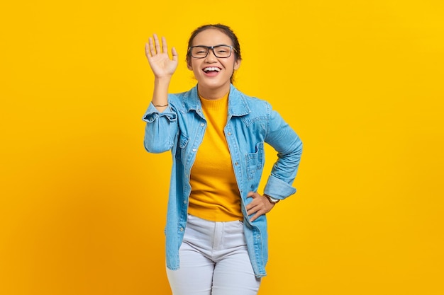Portrait d'une jeune étudiante asiatique gaie dans des vêtements en denim agitant la main à la caméra isolée sur fond jaune. Concept de mode de vie des émotions sincères des gens