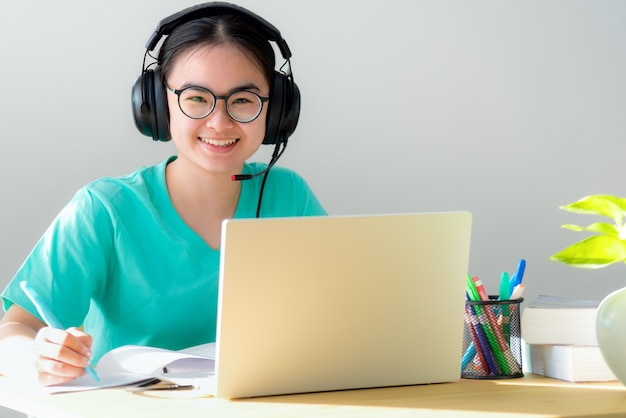 Portrait jeune étudiante asiatique avec des lunettes casque sourire en levant une note sur le livre une adolescente est heureuse d'étudier à distance sur Internet à l'université classe en ligne sur un ordinateur portable à la maison