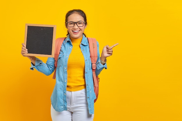 Portrait d'une jeune étudiante asiatique souriante en vêtements décontractés avec sac à dos tenant un tableau blanc et pointant vers l'espace de copie avec le doigt isolé sur fond jaune