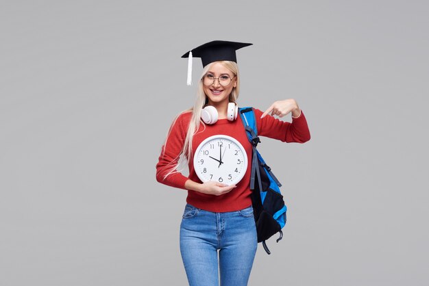 Portrait de jeune étudiante blonde excitée en casquette de diplômé avec sac à dos tenant un gros réveil isolé sur un espace gris. Éducation au collège. Copiez l'espace pour le texte