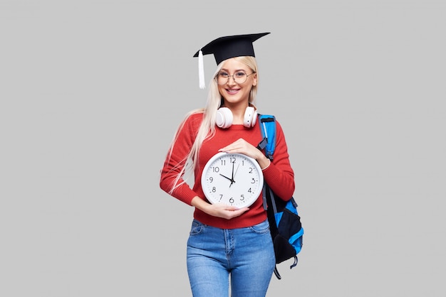 Portrait de jeune étudiante blonde excitée en casquette de diplômé avec sac à dos tenant un gros réveil isolé sur un espace gris. Éducation au collège. Copiez l'espace pour le texte