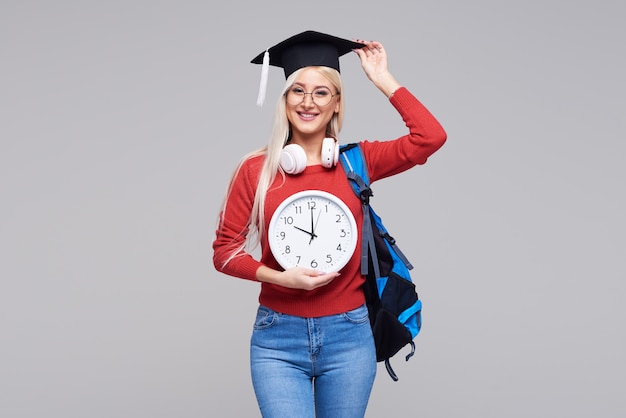 Portrait de jeune étudiante blonde excitée en casquette de diplômé avec sac à dos tenant un gros réveil isolé sur un espace gris. Éducation au collège. Copiez l'espace pour le texte