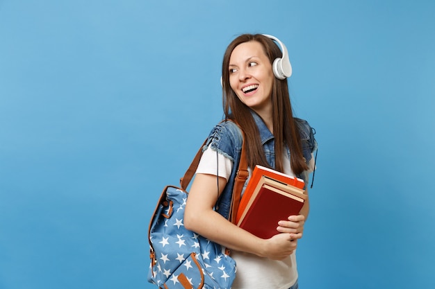 Portrait d'une jeune étudiante curieuse avec un casque à dos écoutant de la musique tenir des livres scolaires en regardant de côté sur un espace de copie isolé sur fond bleu. Éducation au collège universitaire secondaire.