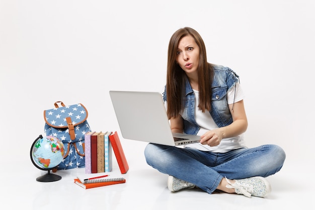 Portrait d'une jeune étudiante décontractée et inquiète tenant un ordinateur portable assis près du globe, sac à dos, livres scolaires isolés