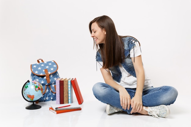 Portrait d'une jeune étudiante décontractée et intéressée dans des vêtements en denim assis, regardant sur un globe, un sac à dos, des livres scolaires isolés