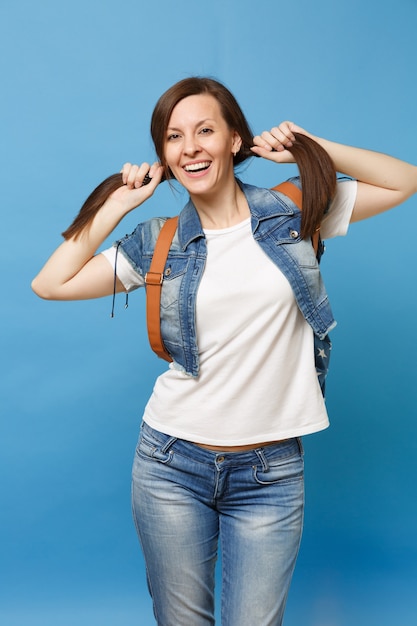 Portrait de jeune étudiante drôle assez souriante en t-shirt blanc, vêtements en jean avec sac à dos tenant des queues de cheval isolées sur fond bleu. L'éducation au collège. Copiez l'espace pour la publicité.