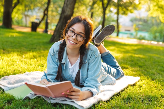 Portrait d'une jeune étudiante heureuse portant des lunettes, assise à l'extérieur dans un parc naturel, écrivant un livre de lecture.