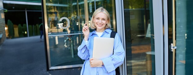 Photo portrait d'une jeune étudiante insouciante avec un ordinateur portable et un sac à dos appuyé sur le bâtiment du campus