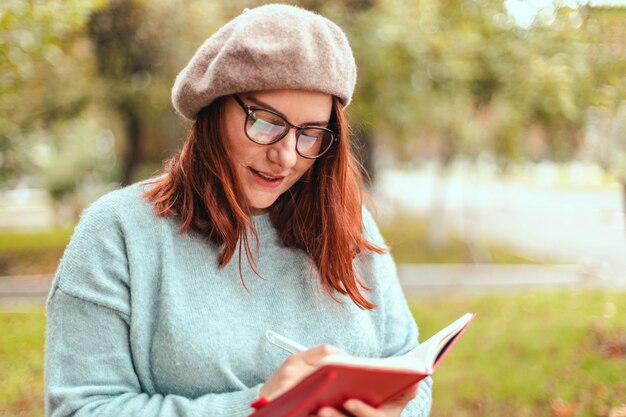 Portrait d'une jeune étudiante joyeuse, écrivant des notes dans un ordinateur portable assis à l'extérieur dans un parc naturel