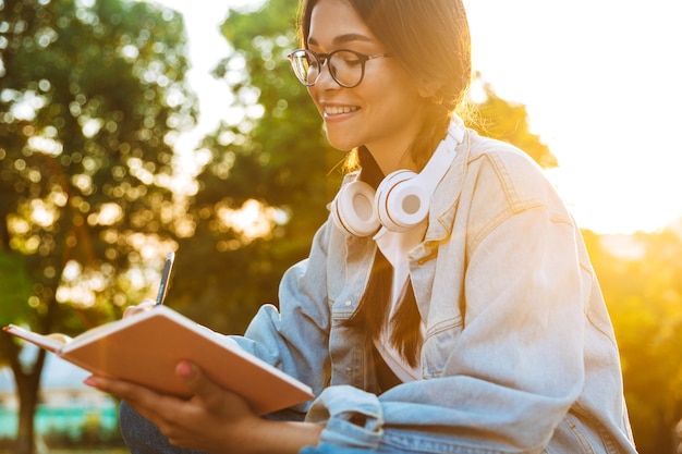 Portrait D'une Jeune étudiante Joyeuse Portant Des Lunettes Assise à L'extérieur Dans Un Parc Naturel écoutant De La Musique Avec Des écouteurs Et écrivant Des Notes