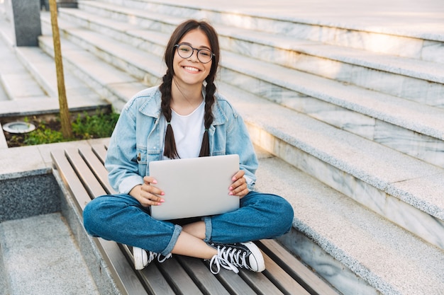 Portrait d'une jeune étudiante mignonne souriante et heureuse portant des lunettes assise sur un banc à l'extérieur dans un parc naturel à l'aide d'un ordinateur portable