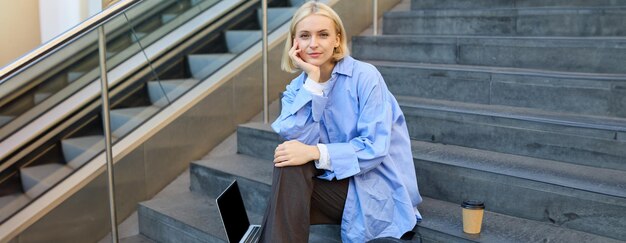 Portrait d'une jeune étudiante moderne ou d'une travailleuse indépendante assise sur les escaliers extérieurs se reposant en ville