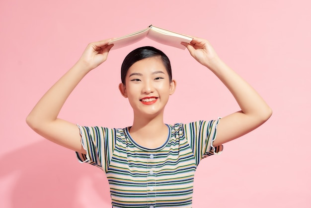 Portrait d'une jeune étudiante souriante drôle avec un livre ouvert sur la tête sur fond rose