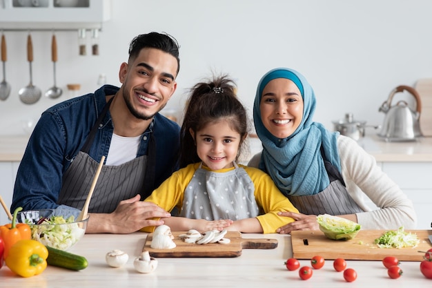 Portrait d'une jeune famille heureuse du Moyen-Orient cuisinant ensemble dans la cuisine, de joyeux parents arabes et leur petite fille en tabliers assis à table tout en préparant des plats savoureux ensemble