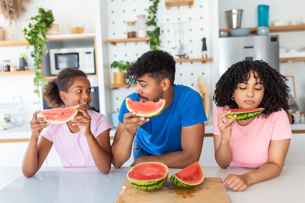 Portrait d'une jeune famille joyeuse prenant une bouchée d'une pastèque Famille afro-américaine debout ensemble dans une cuisine mangeant de la pastèque