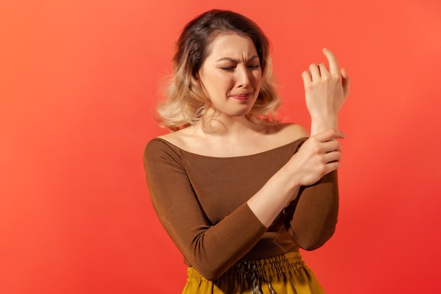 Photo portrait d'une jeune femme adulte bouleversée aux cheveux blonds en chemisier marron debout avec une grimace de douleur massant le poignet endolori souffrant d'une blessure à la main ou d'une entorse prise de vue en studio intérieure isolée sur fond rouge