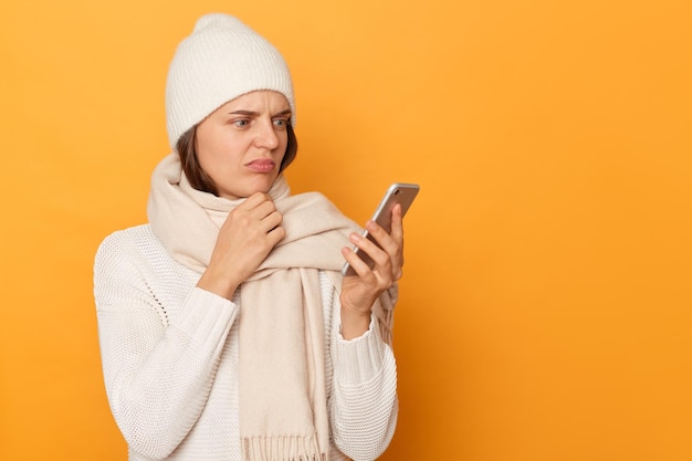 Portrait d'une jeune femme adulte caucasienne perplexe et confuse portant un pull blanc, un chapeau et une écharpe posant isolé sur fond jaune à l'aide d'un téléphone portable tenant le menton ne comprend pas quelque chose