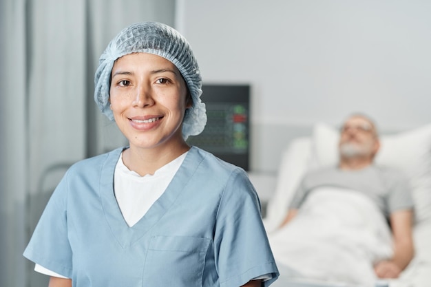 Portrait d'une jeune femme adulte confiante travaillant dans un hôpital souriant à la caméra, son patient allongé dans le lit en arrière-plan