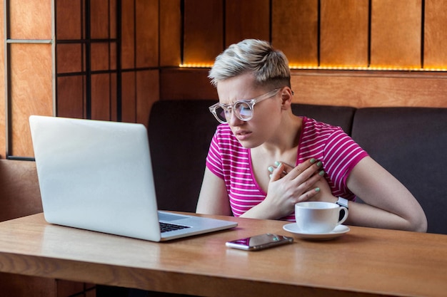 Portrait d'une jeune femme adulte malade aux cheveux blonds courts en t-shirt rose et lunettes assise dans un café et touchant le cœur avec la main a une douleur intense blessée infarctus respiratoire Soins de santé à l'intérieur